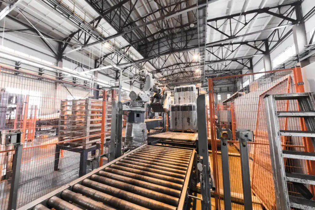 Zautomatyzowana linia produkcyjna w fabryce, wyposażona w przenośnik rolkowy, maszyny przemysłowe oraz pomarańczowe ogrodzenia ochronne, usytuowana pod wysokim stropem ze stalowymi kratownicami.